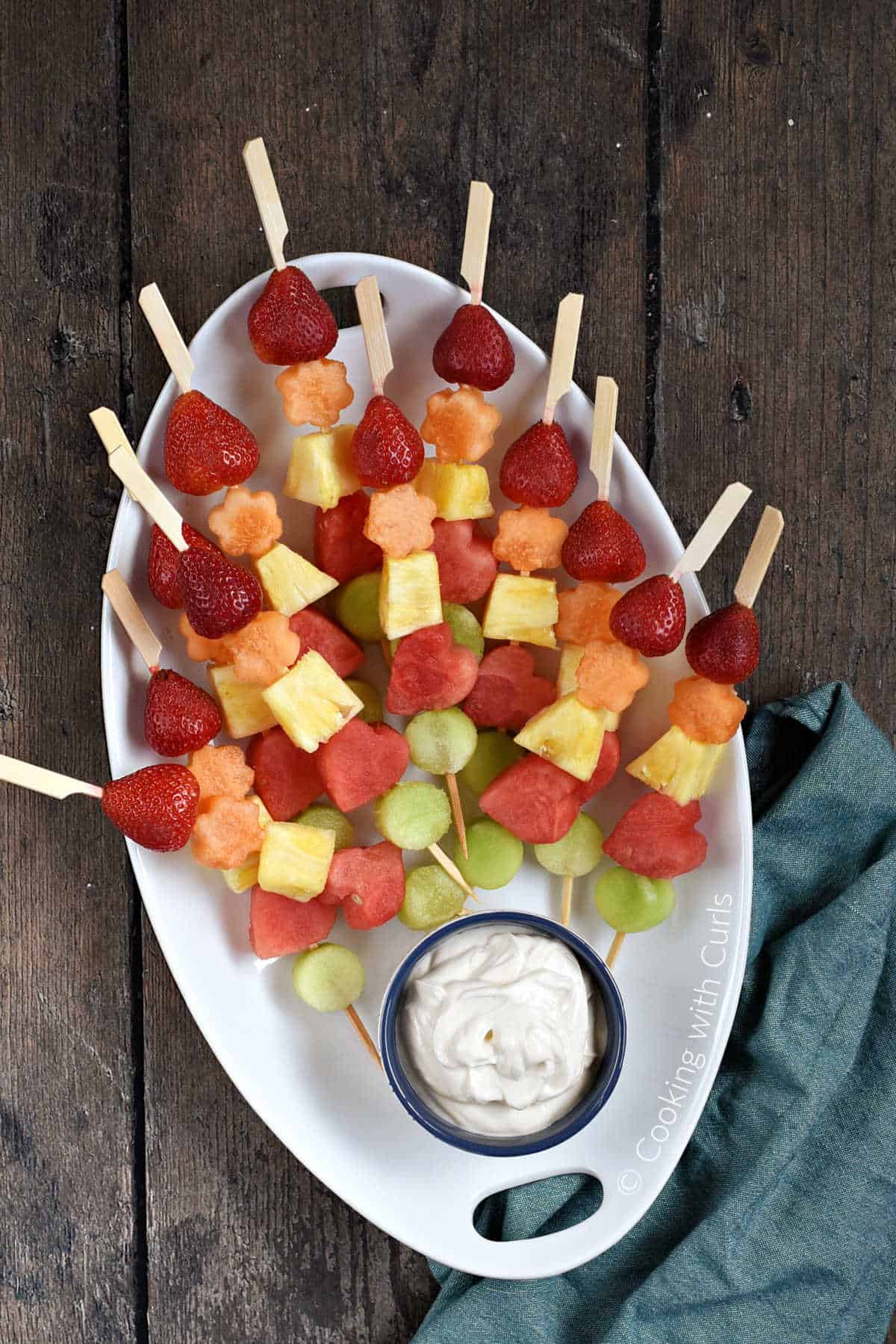 Fresh fruits threaded onto wood skewers, stacked on an oval platter with a bowl of yogurt dip at the bottom.