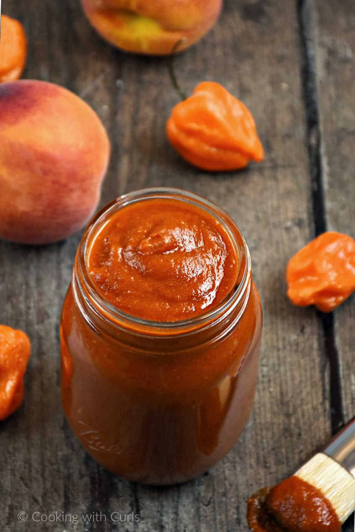 Peach barbecue sauce in a mason jar surrounded by fresh peaches and peppers.
