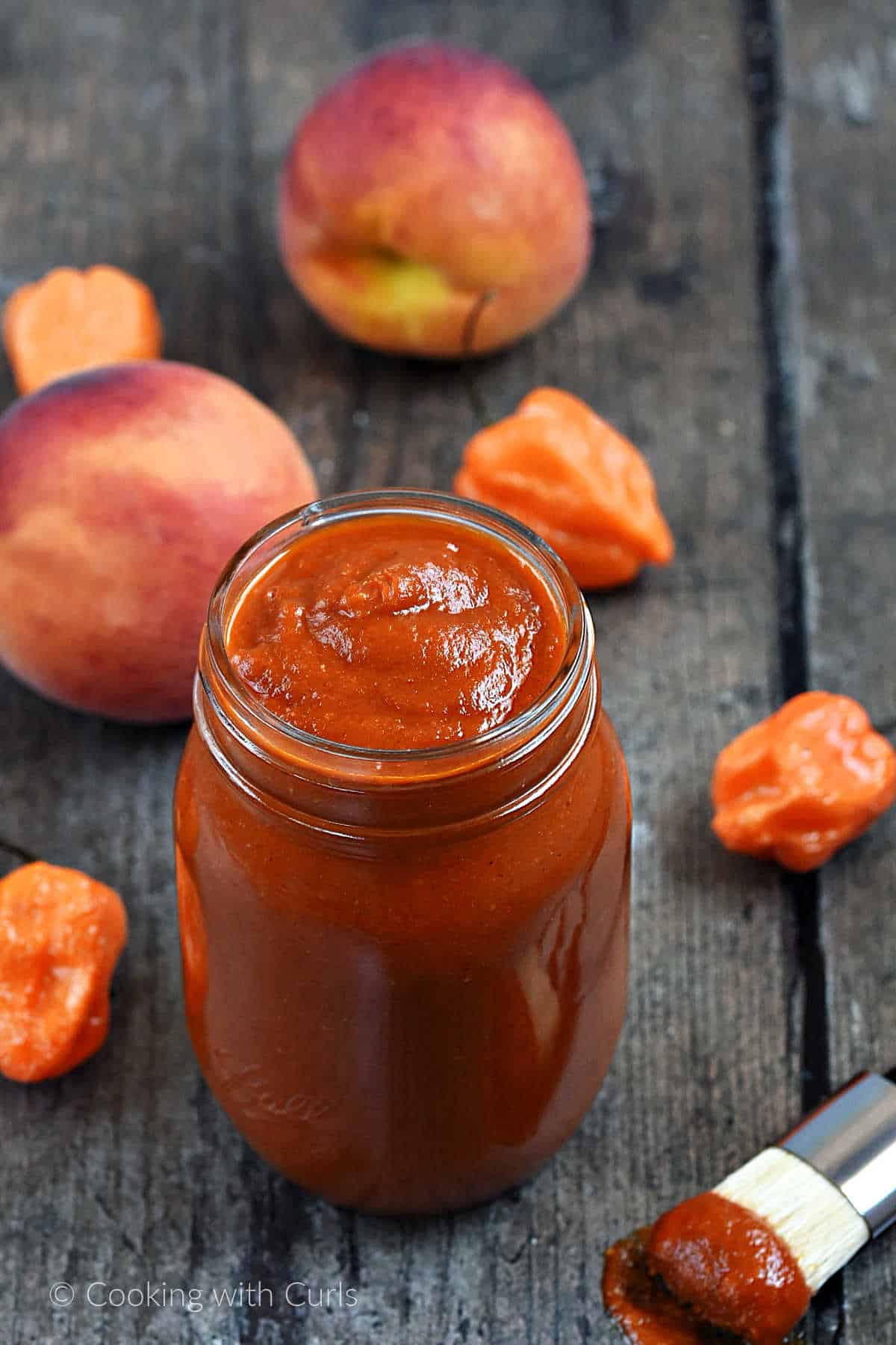Mason jar filled with peach habanero bbq sauce surrounded by fresh peaches and peppers.