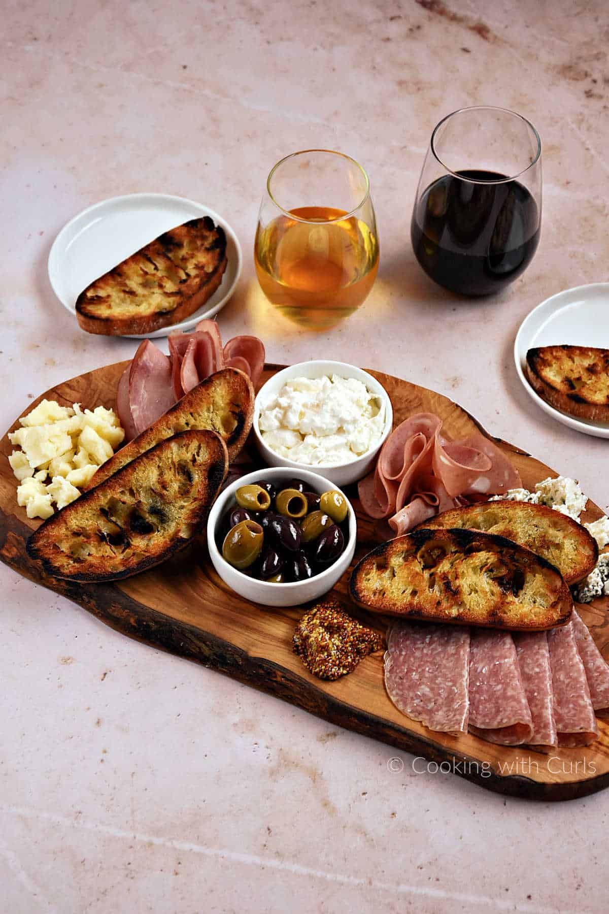 Italian deli meats and cheeses on a wood board with olives, mustard, and grilled bread.