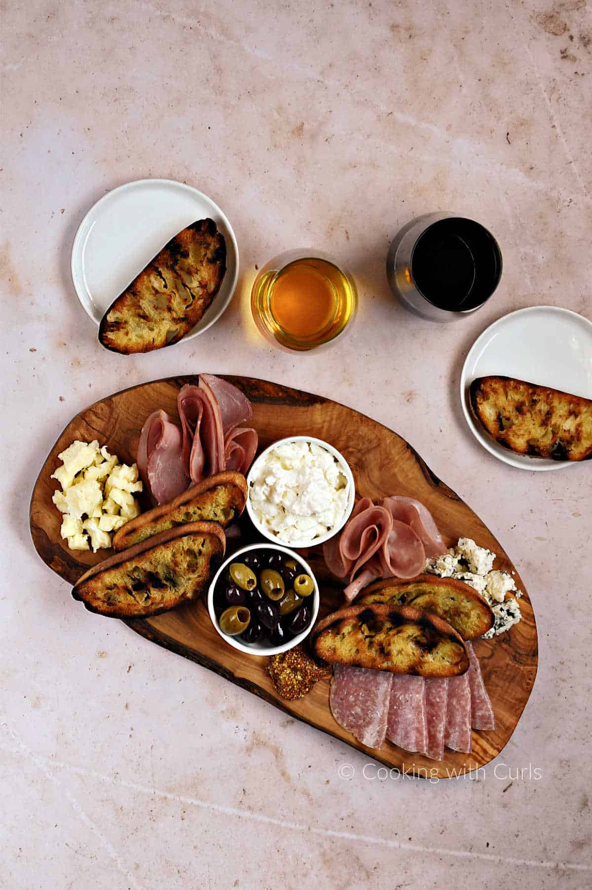 Looking down on a wood board topped with Italian meats and cheese, olives, and grilled bread with glasses of wine in the background.