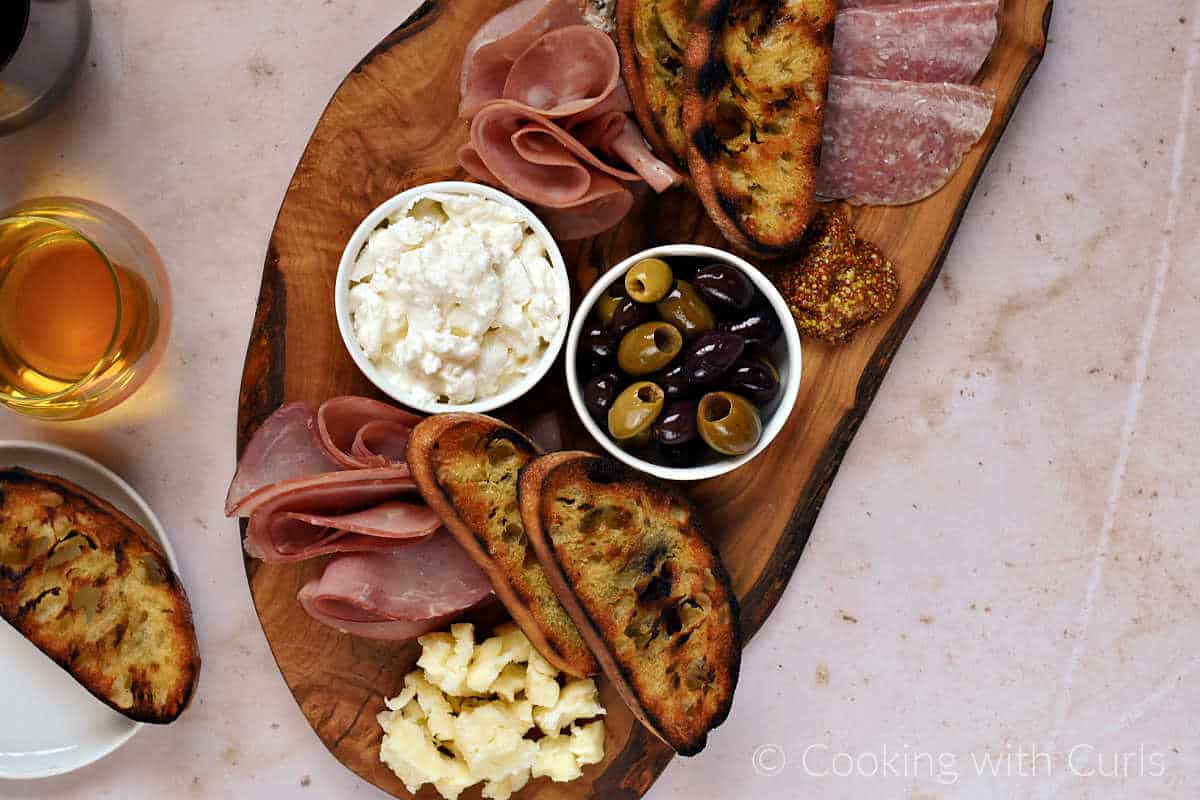 Horizontal image of wood board topped with Italian meats and cheeses with a glass of wine on the side.