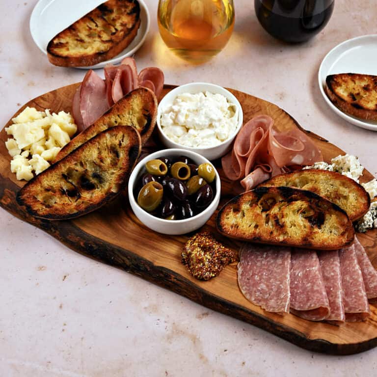Italian deli meats and cheeses on a wood board with olives, mustard, and grilled bread.