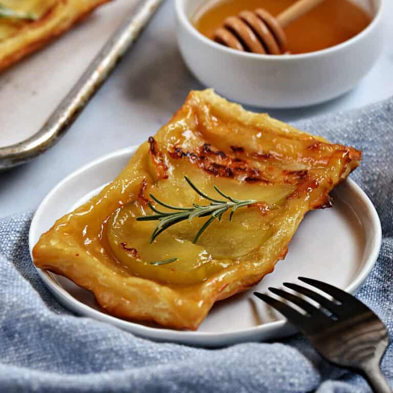 An apple tart with puff pastry crust topped with a sprig of fresh rosemary with a bowl a honey in the background.