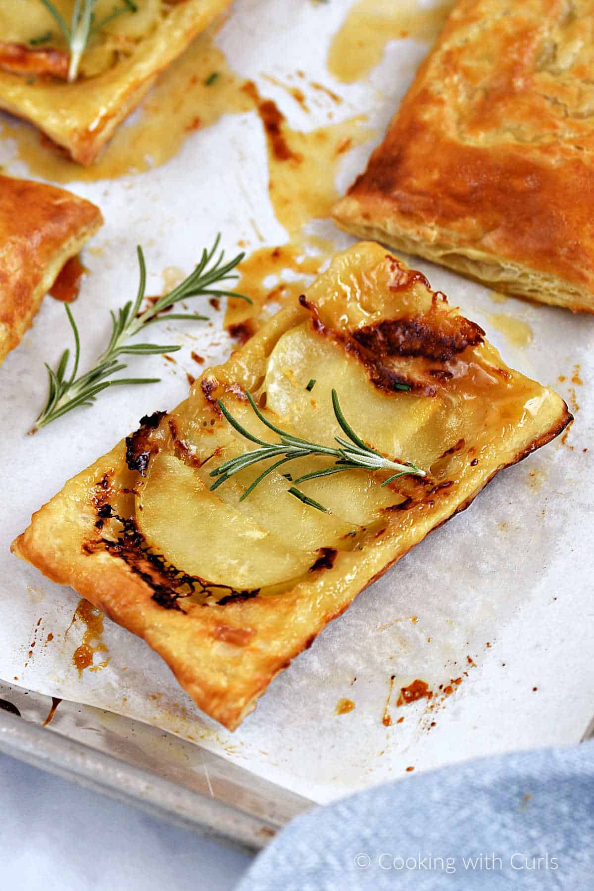 A puff pastry apple tart topped with a sprig of fresh rosemary sitting on a parchment lined baking sheet surrounded by three other tarts.