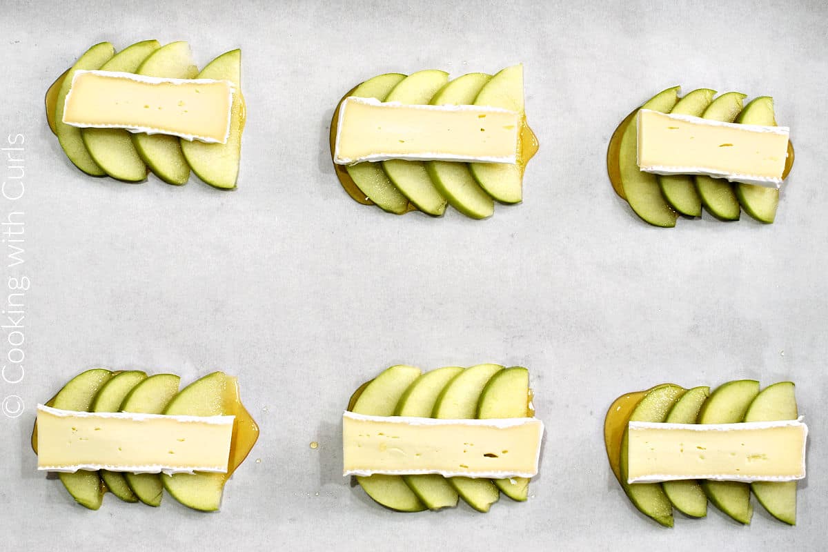 Apple slices topped with a slice of brie arranged in six spaces on a parchment lined baking sheet.