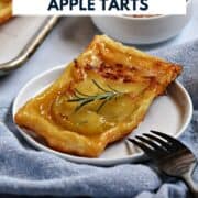 An apple tart with puff pastry crust topped with a sprig of fresh rosemary with a bowl a honey and a tray of tarts in the background and title graphic across the top.