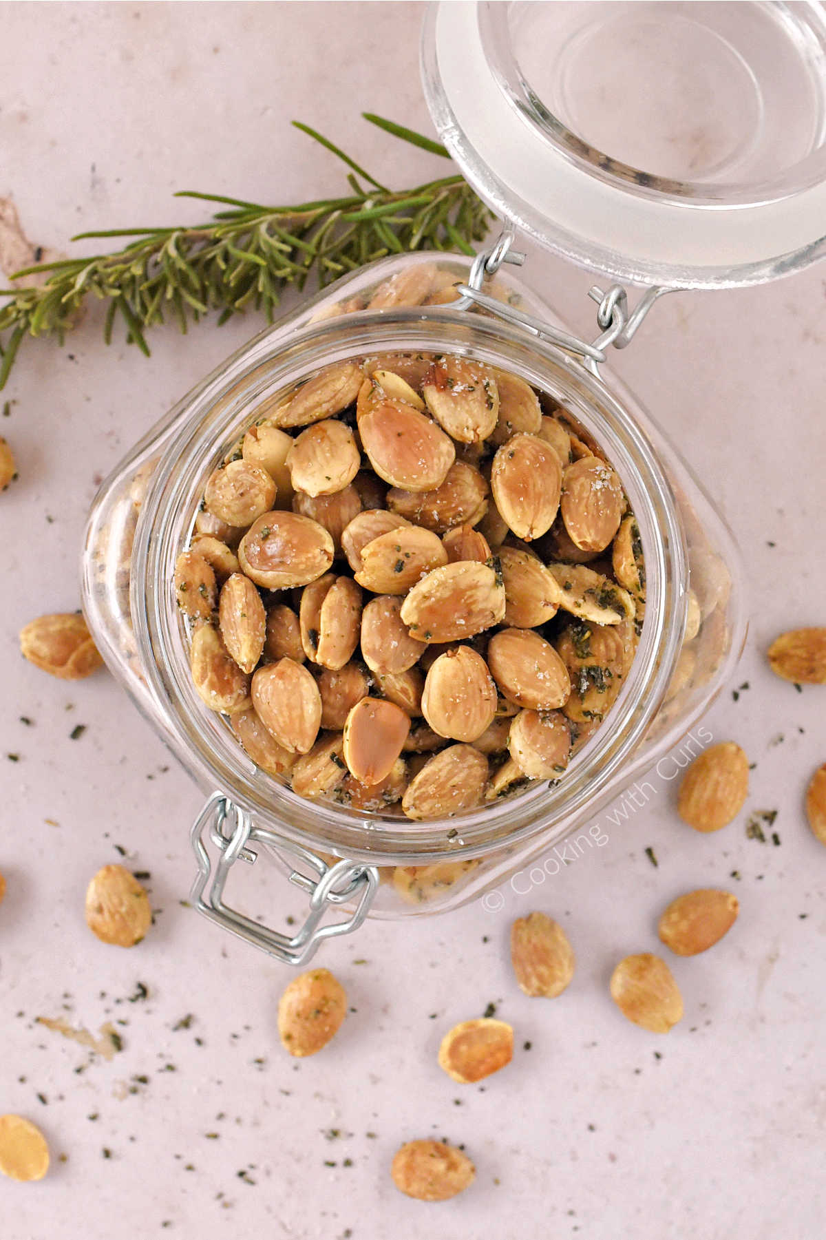 Looking down on a jar filled with rosemary marcona almonds surrounded by almonds and a sprig of rosemary.
