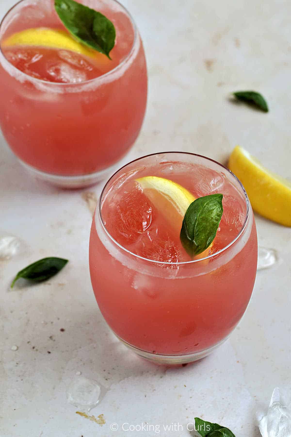 Two glasses of pink watermelon basil lemonade drinks with a lemon wedge and basil garnish.