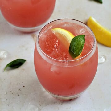 Two glasses of pink watermelon basil lemonade drinks with a lemon wedge and basil garnish.