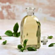 Basil simple syrup in a glass bottle surrounded by basil leaves on a pink background.