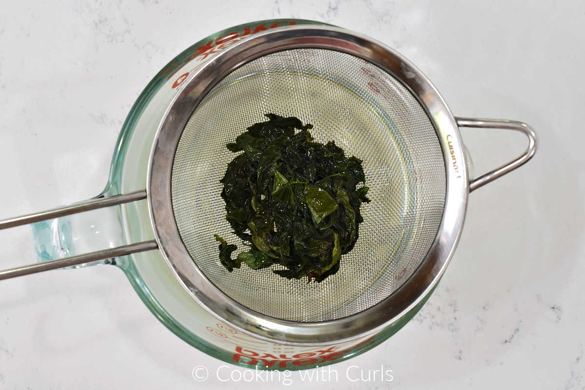 Cooked basil leaves in a strainer over a measuring cup.