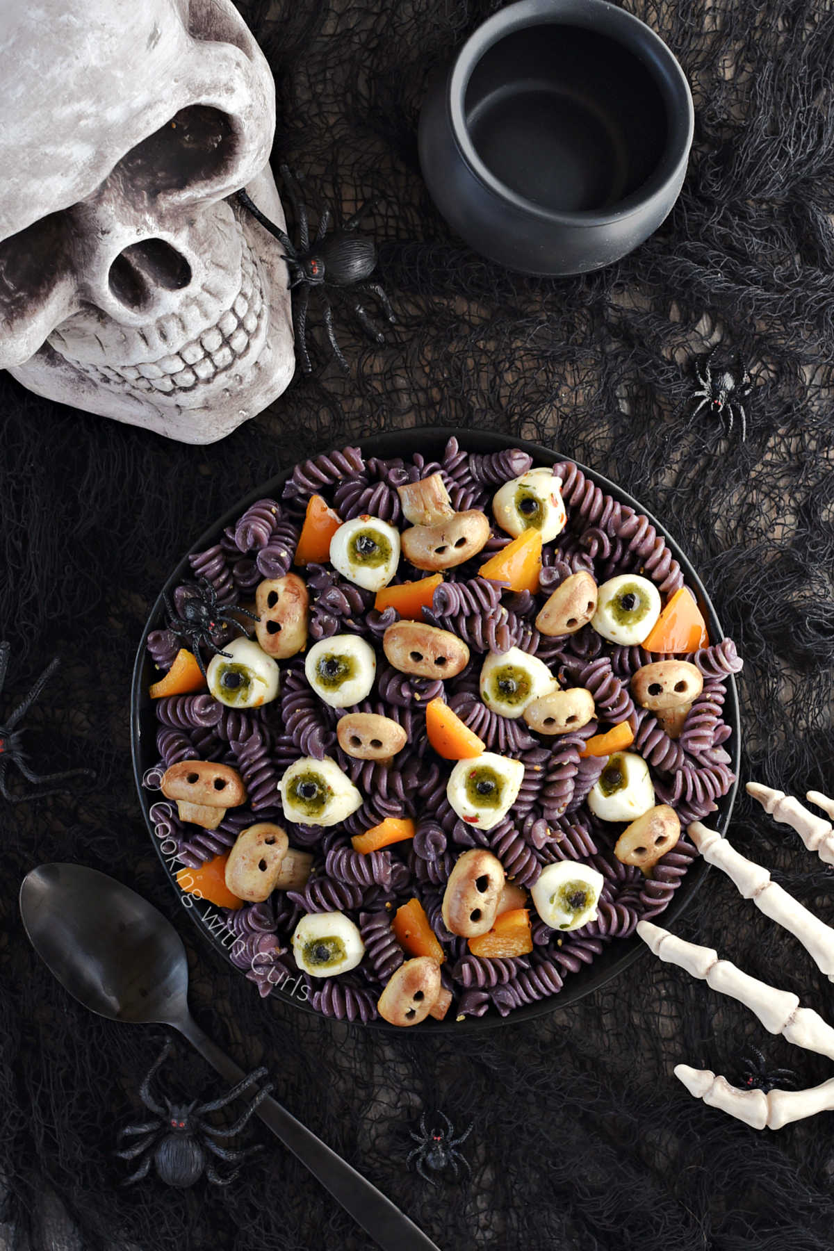 Black spiral pasta, mushroom skulls, mozzarella eyeballs, and orange triangles in a black serving bowl with skull in the background and skeleton hand on the edge of the bowl.