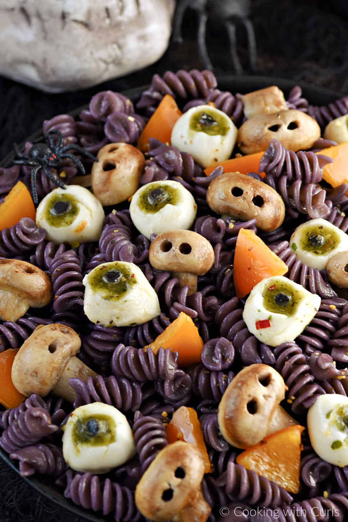 Black spiral pasta, mushroom skulls, mozzarella eyeballs, and orange triangles in a black serving bowl.