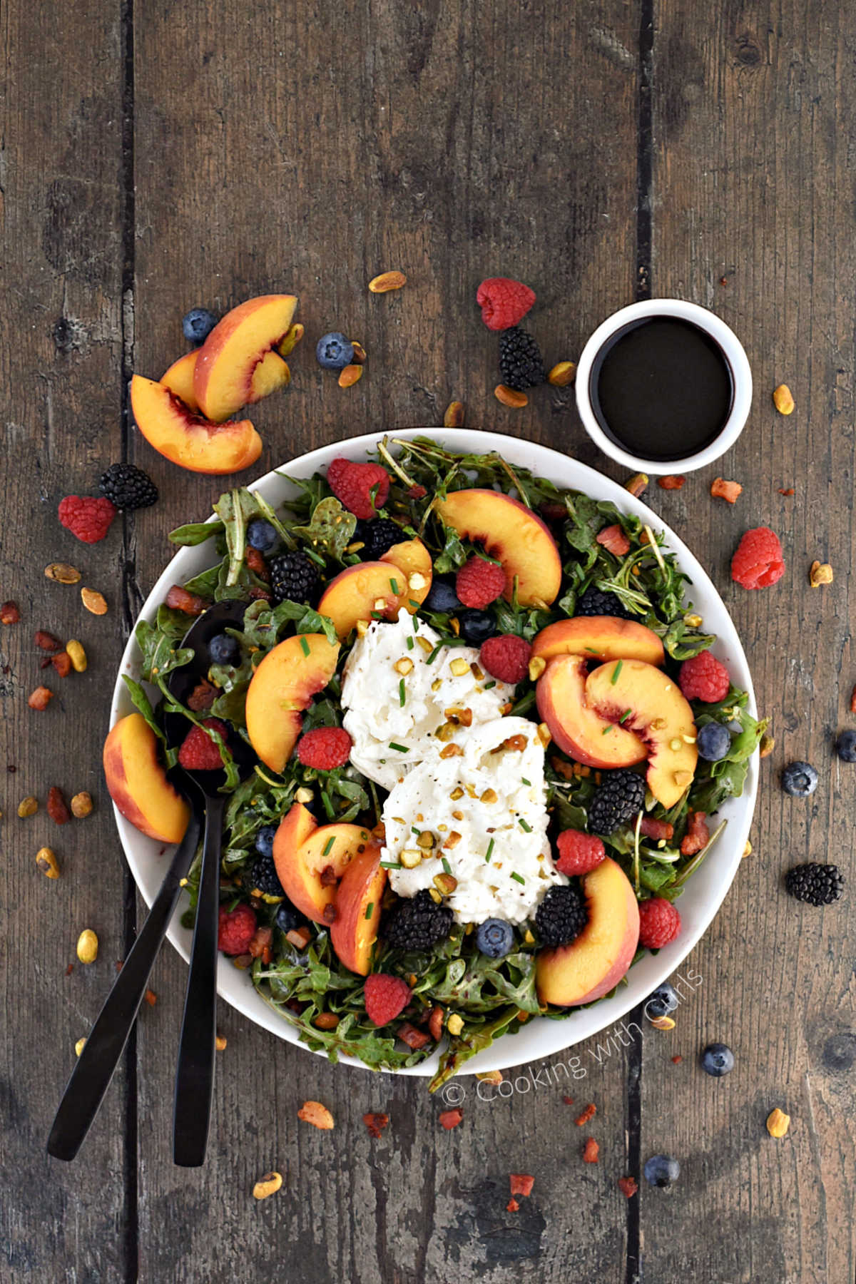 Sliced peaches, fresh berries, and fresh burrata on a bed of arugula with additional ingredients scattered around the platter.
