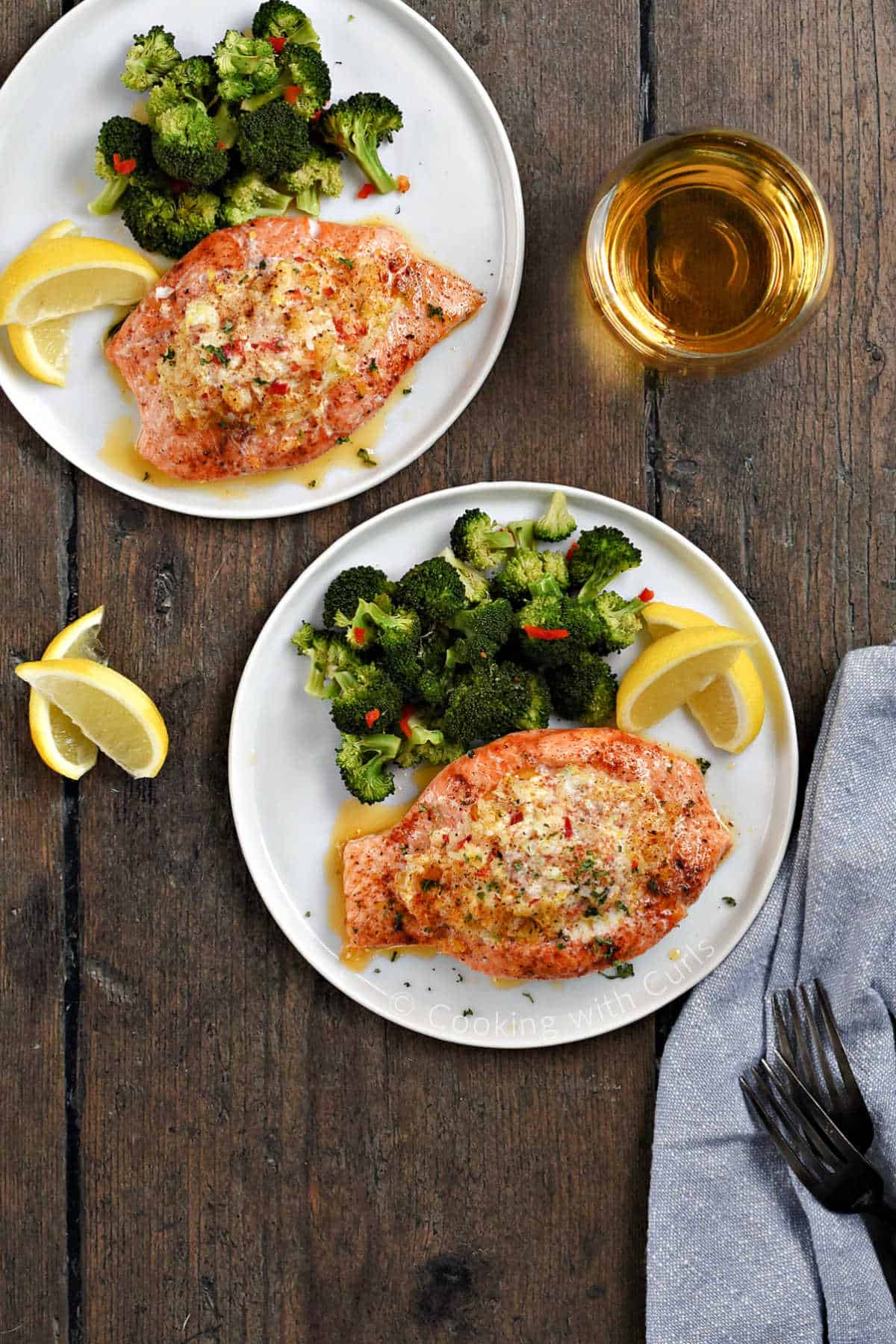 Two plates with crab stuffed salmon, broccoli and lemon wedges with a glass of white wine in the background.