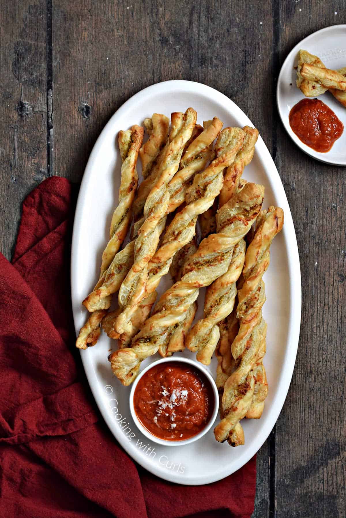 Pesto puff pastry twists stacked on a large platter with a small bowl of marinara on the side.