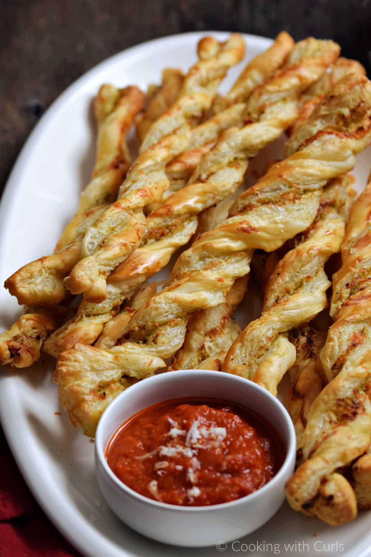 Pesto puff pastry twists on a large platter with a side of marinara sauce.