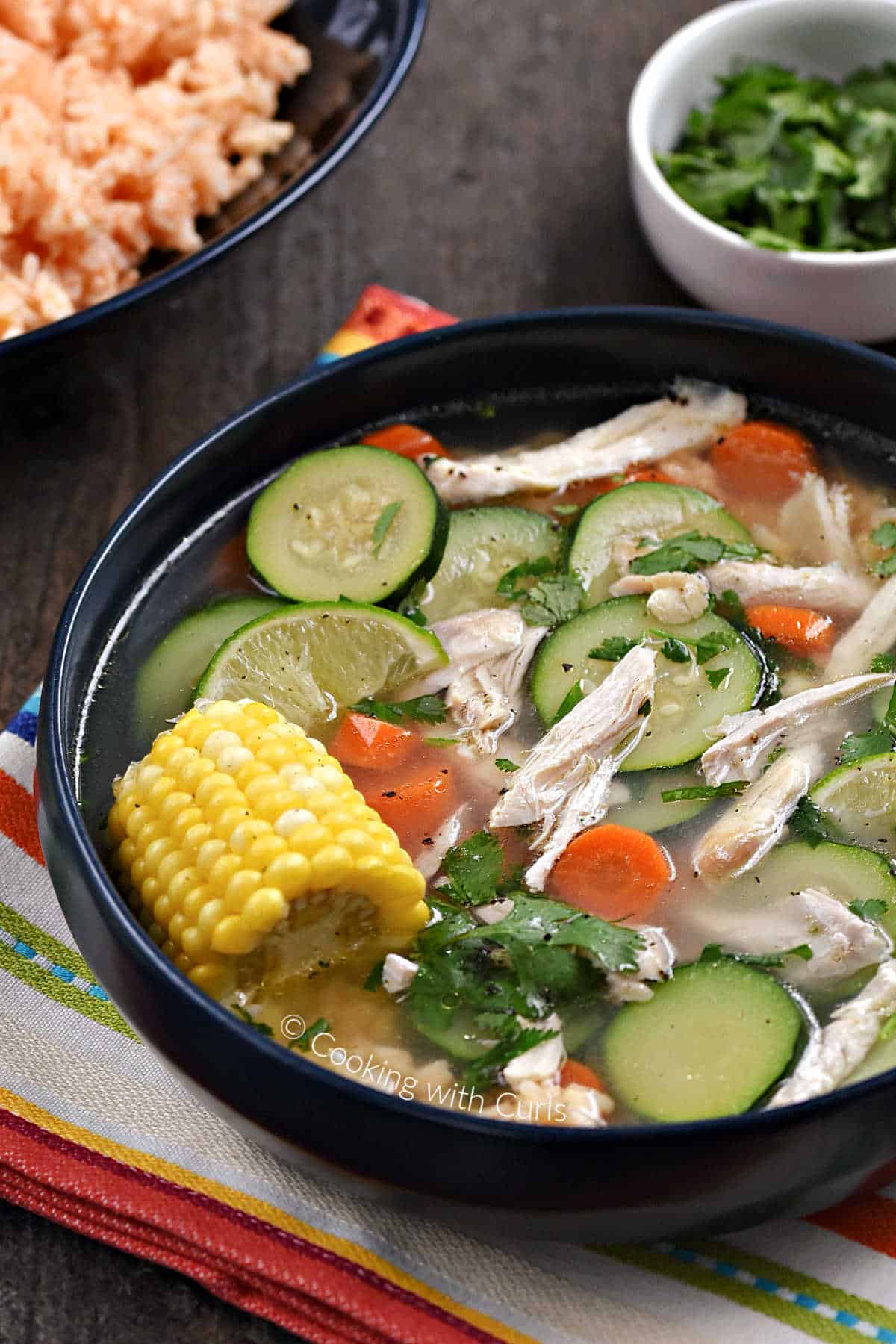 Chicken, sliced zucchini, carrots, and corn on the cob in a bowl with bowls of rice and cilantro in the background.