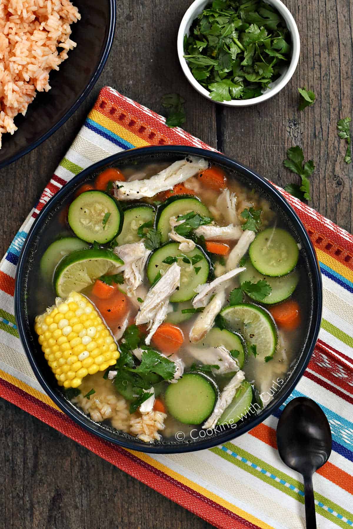 Chicken, sliced zucchini, carrots, and corn on the cob in a bowl with bowls of rice and cilantro in the background.