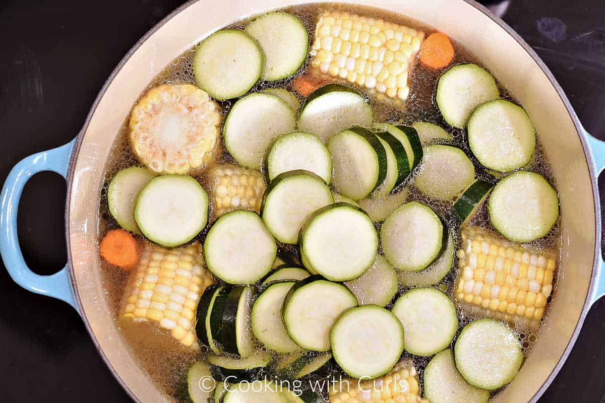 Sliced zucchini, carrots, and corn on the cob with chicken broth in a large pot.