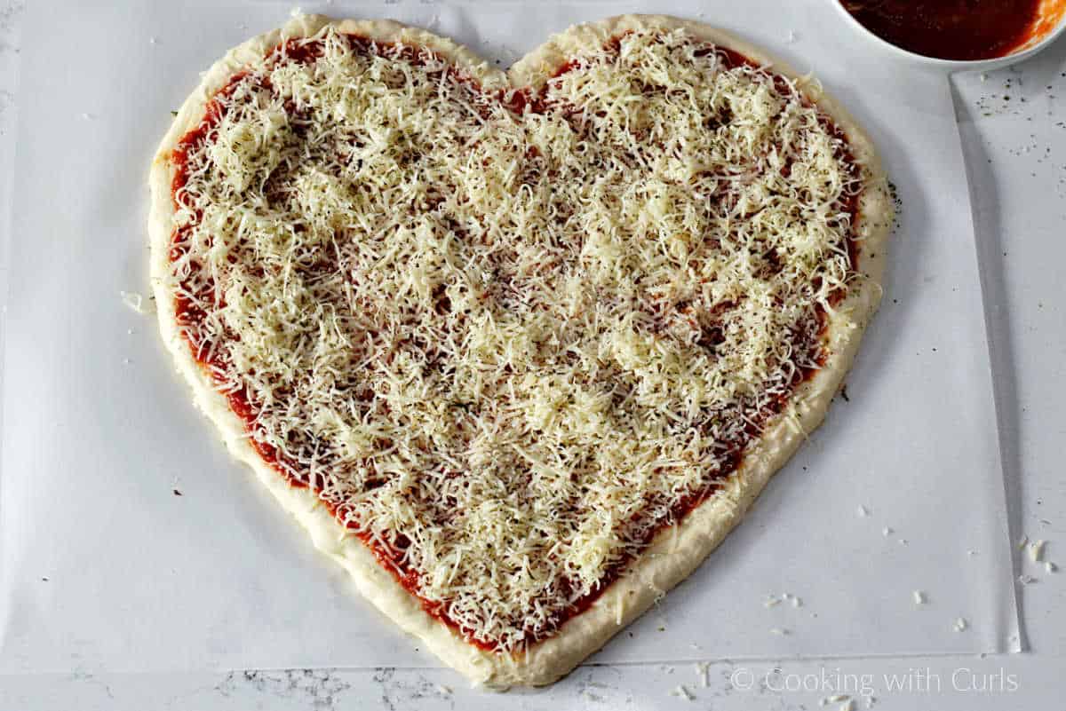 Heart shaped pizza dough topped with red sauce, shredded mozzarella and parmesan cheese.