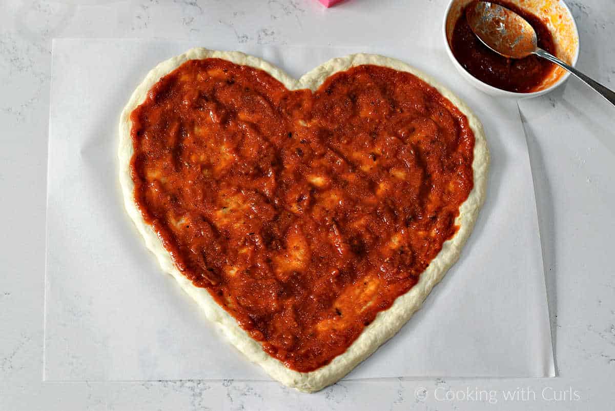 Heart shaped pizza crust topped with red pizza sauce.