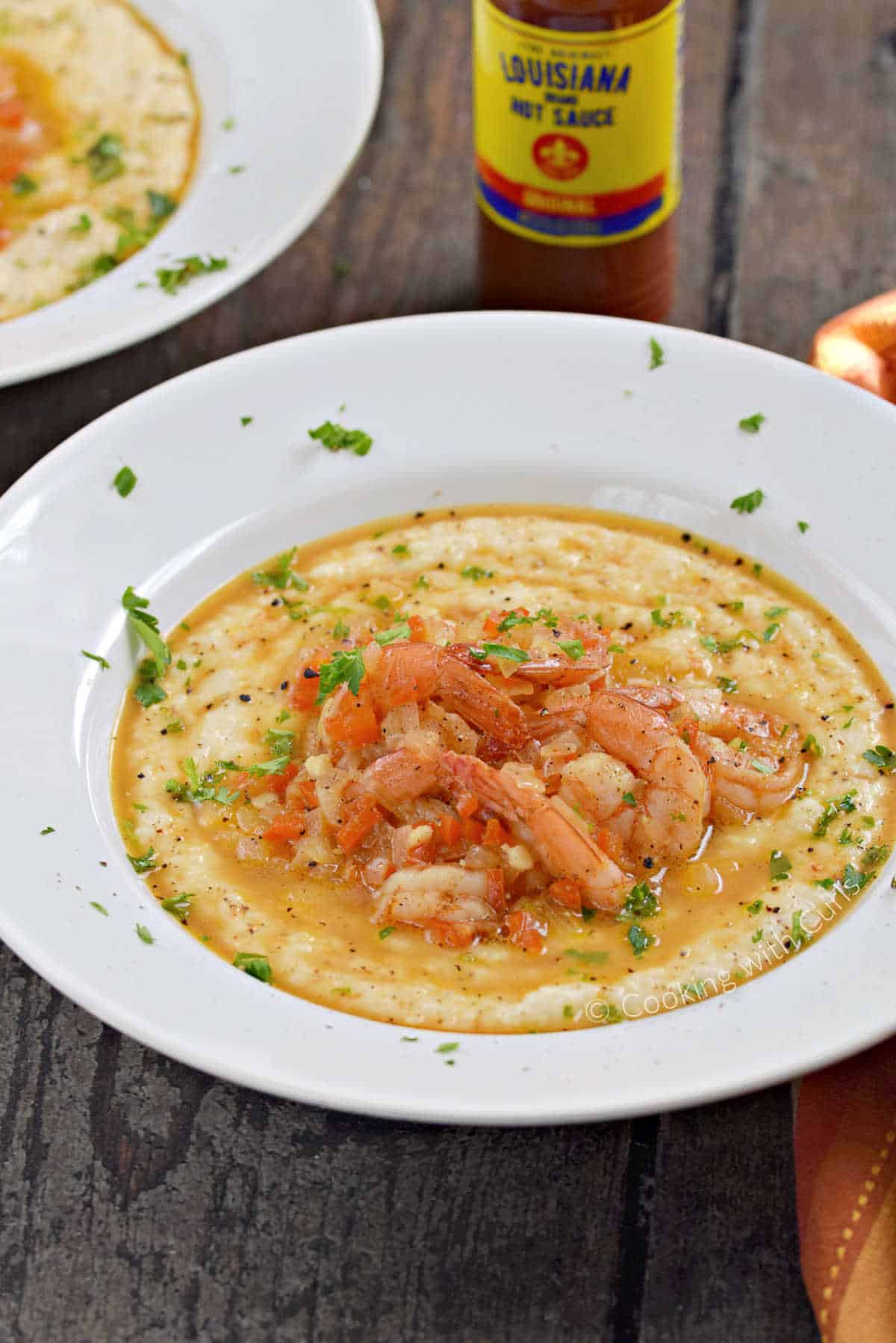 Two bowls of cheesy grits topped with seasoned shrimp and parsley with a bottle of hot sauce between them.