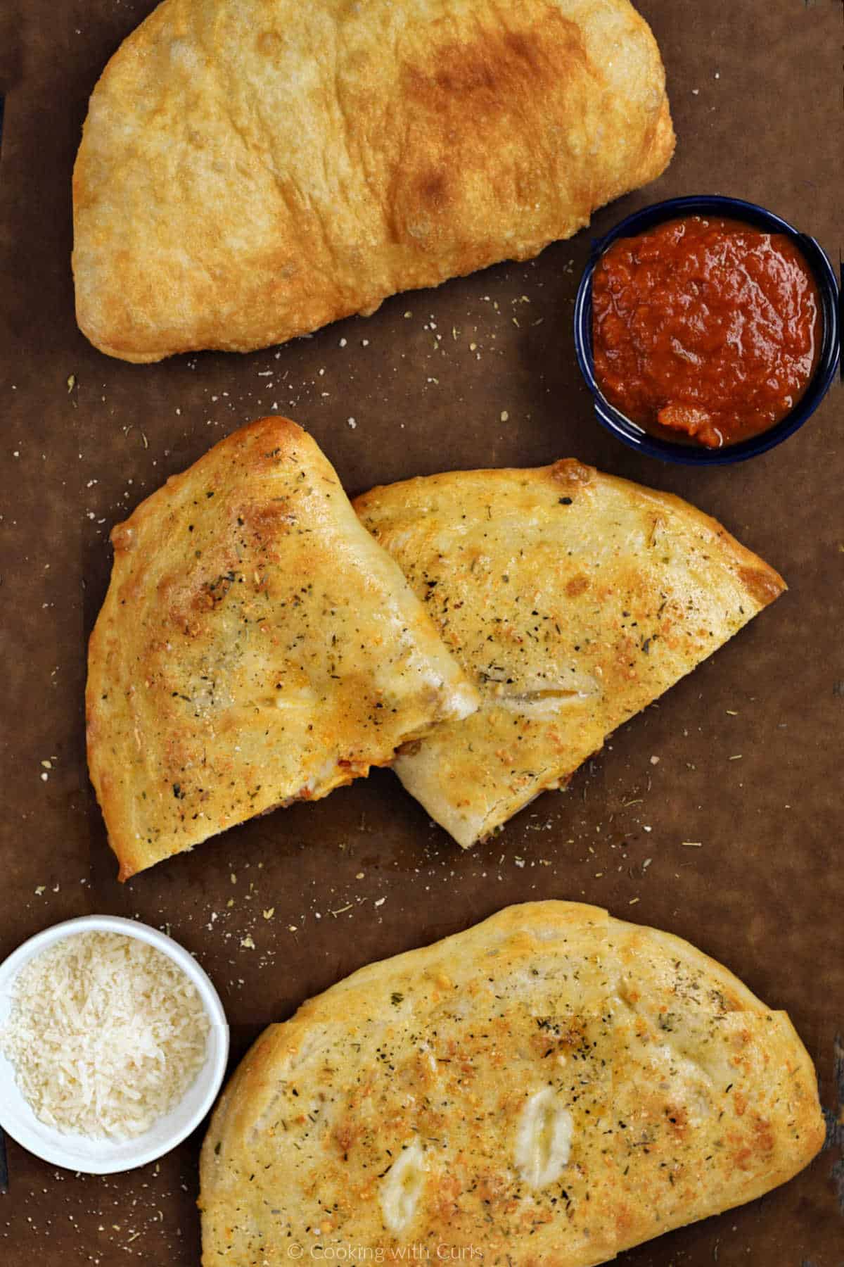 Two whole calzones and one cut in half on a board with pizza sauce in bowl.
