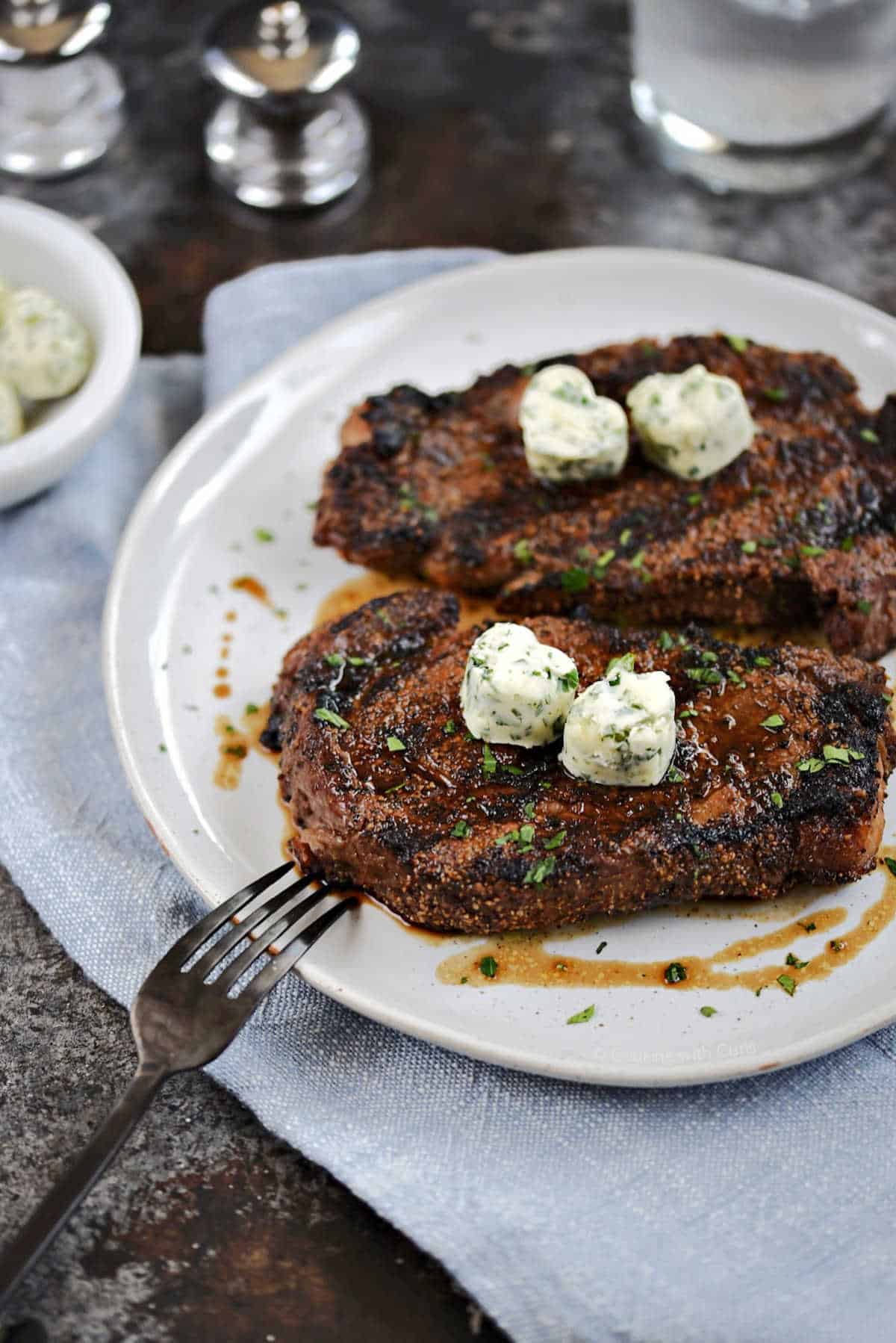 Two perfectly grilled ribeye steaks with hart shaped herb butter on top.