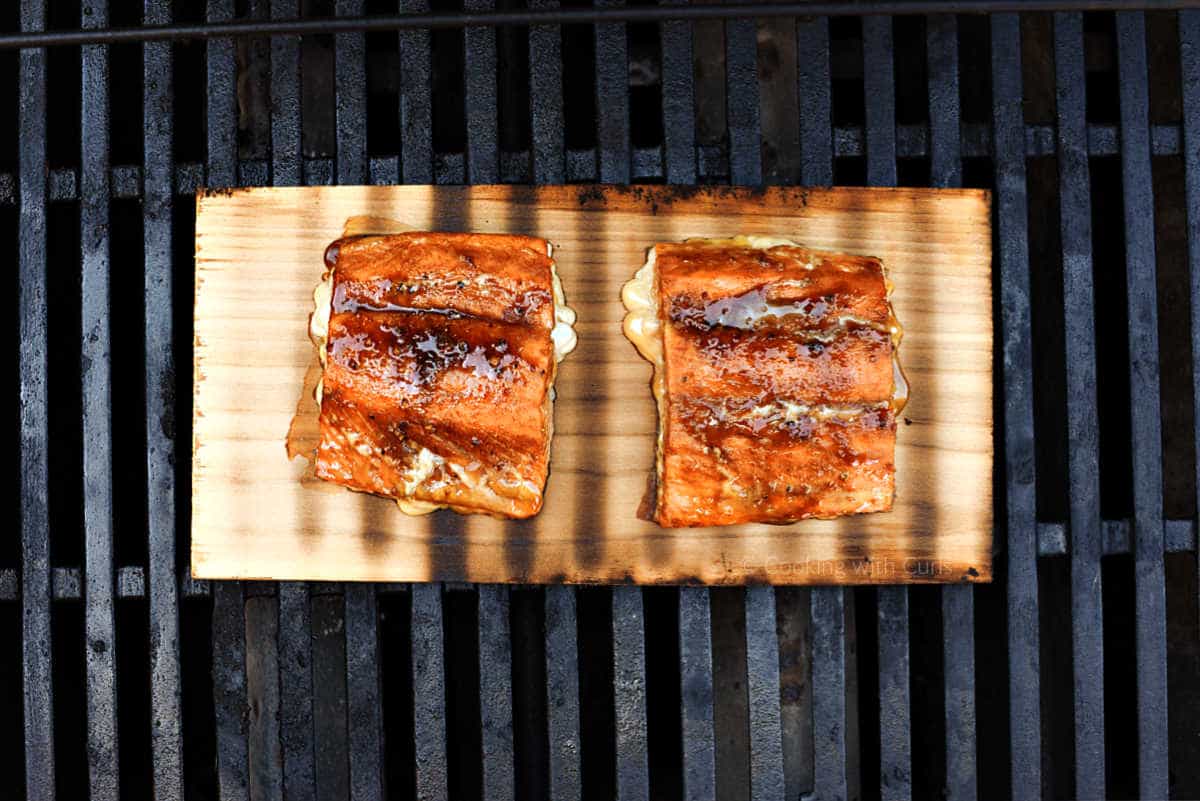 Two maple glazed salmon filets on a cedar plank sitting on grill grates.
