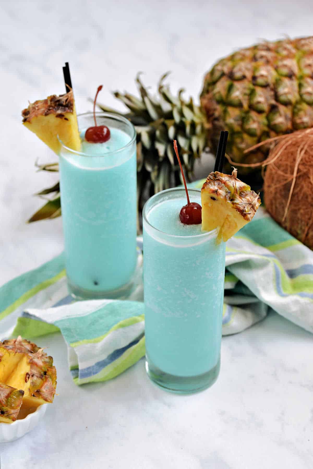 Two frozen blue cocktails in highball glasses with pineapple wedges and cherries.