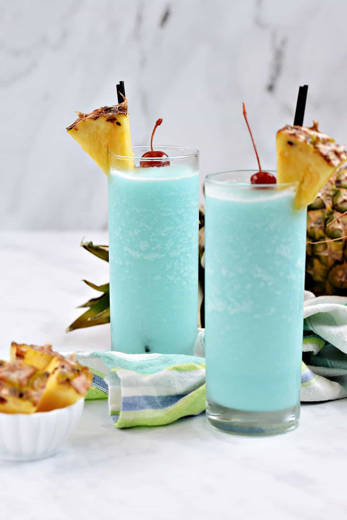 Two frozen Blue Hawaiian cocktails in tall glasses with pineapple wedges and cherry garnish.