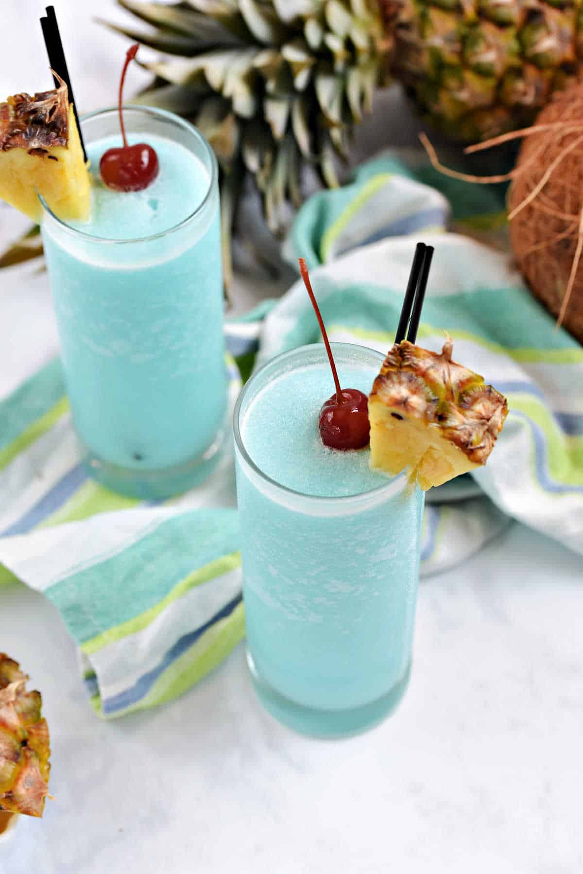 Two glasses with frosty blue Hawaiian cocktails garnished with pineapple wedges and a cherry.