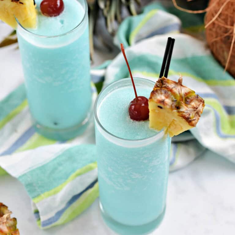 Two glasses with frosty blue Hawaiian cocktails garnished with pineapple wedges and a cherry.