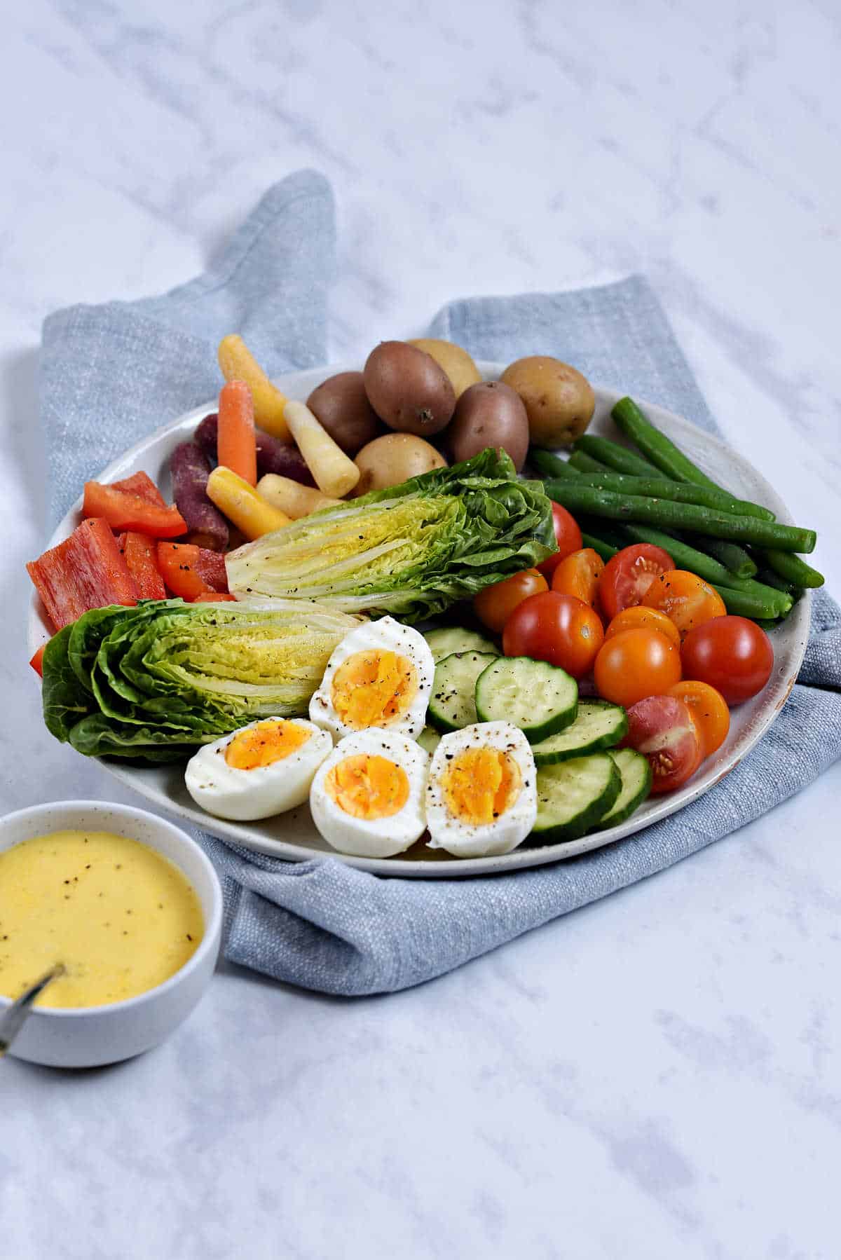 A platter filled with green beans, potatoes, hard boiled eggs, and other seasonal vegetables and a small bowl of aioli.