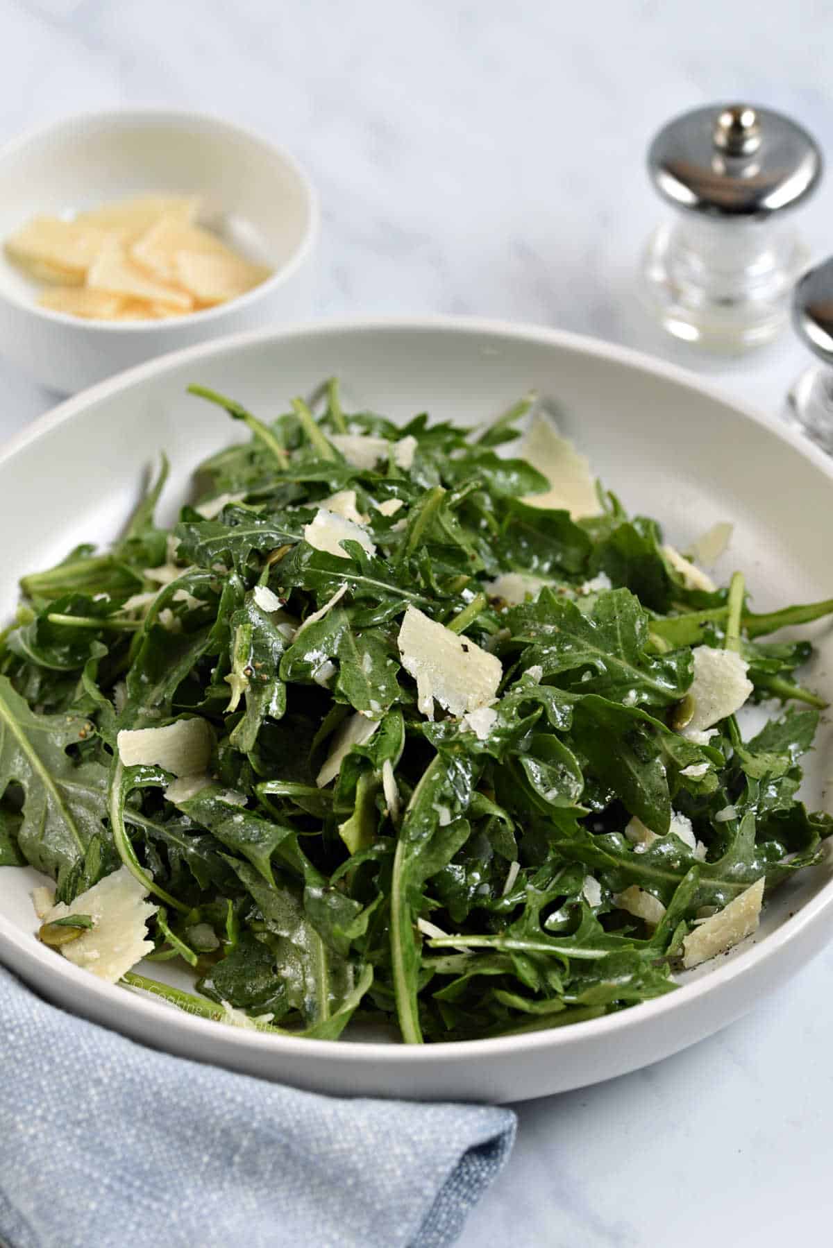 Arugula salad tossed with shaved parmesan cheese, herbs, and pumpkin seeds with bowl of parmesan in the background.