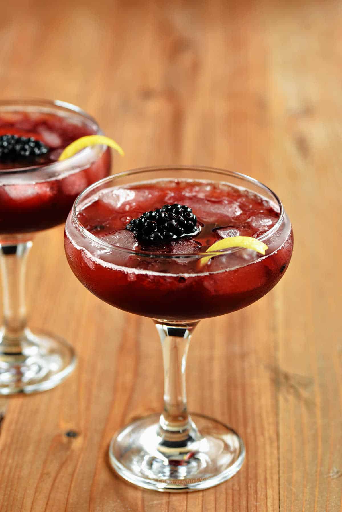 Two coupe glasses with a rich, red drink, ice cubes, a blackberry, and lemon twist.