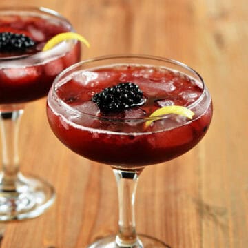 Two coupe glasses with a rich, red drink, ice cubes, a blackberry, and lemon twist.