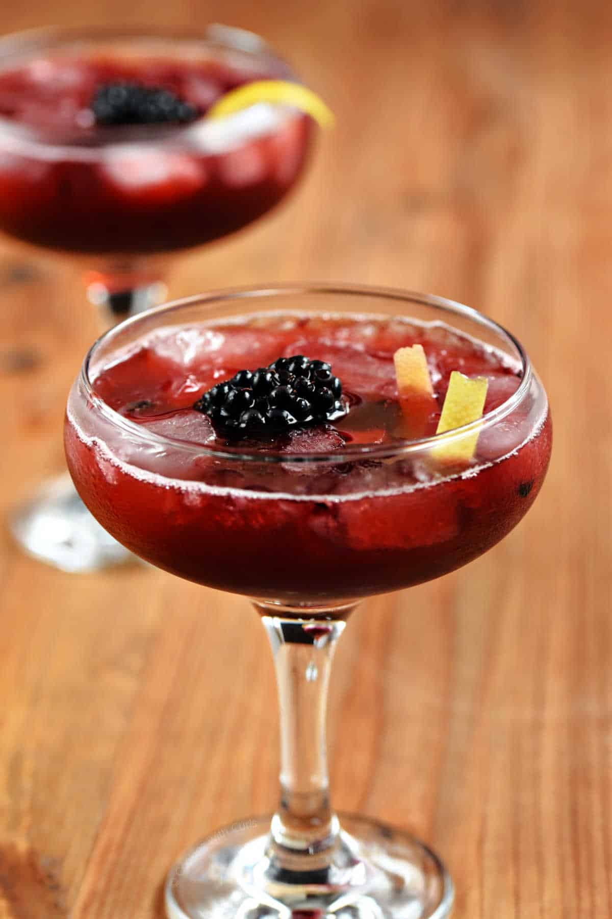 Two coupe glasses with a rich, red drink, ice cubes, a blackberry, and lemon twist.