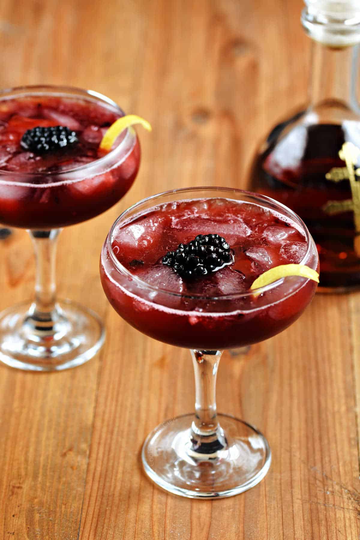 Two coupe glasses with a rich, red drink, ice cubes, a blackberry, and lemon twist with a bottle of cognac in the background.