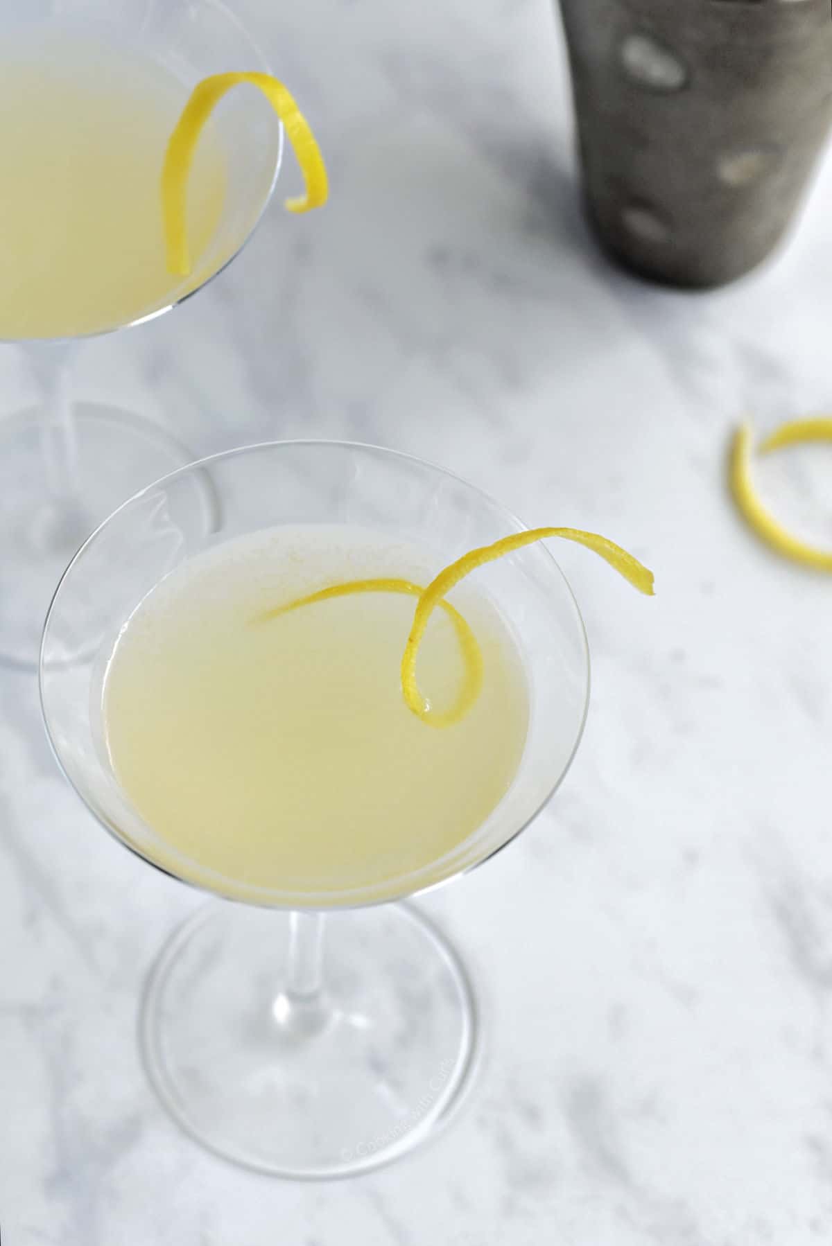 Two yellow cocktails with lemon twist and cocktail shaker in the background.