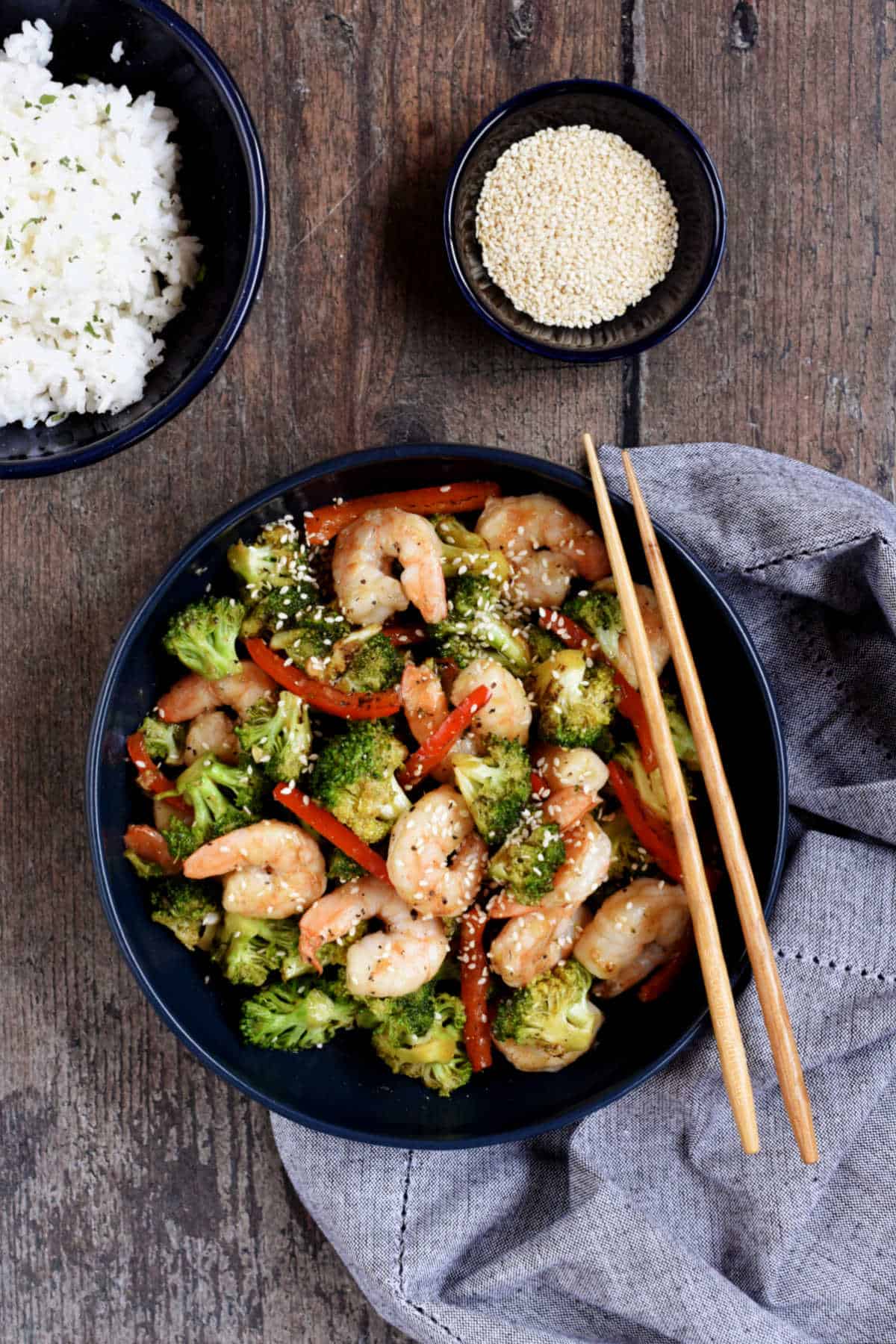 Stir fry shrimp, broccoli, red pepper strips, and toasted sesame seeds in a bowl with chop sticks on the edge.