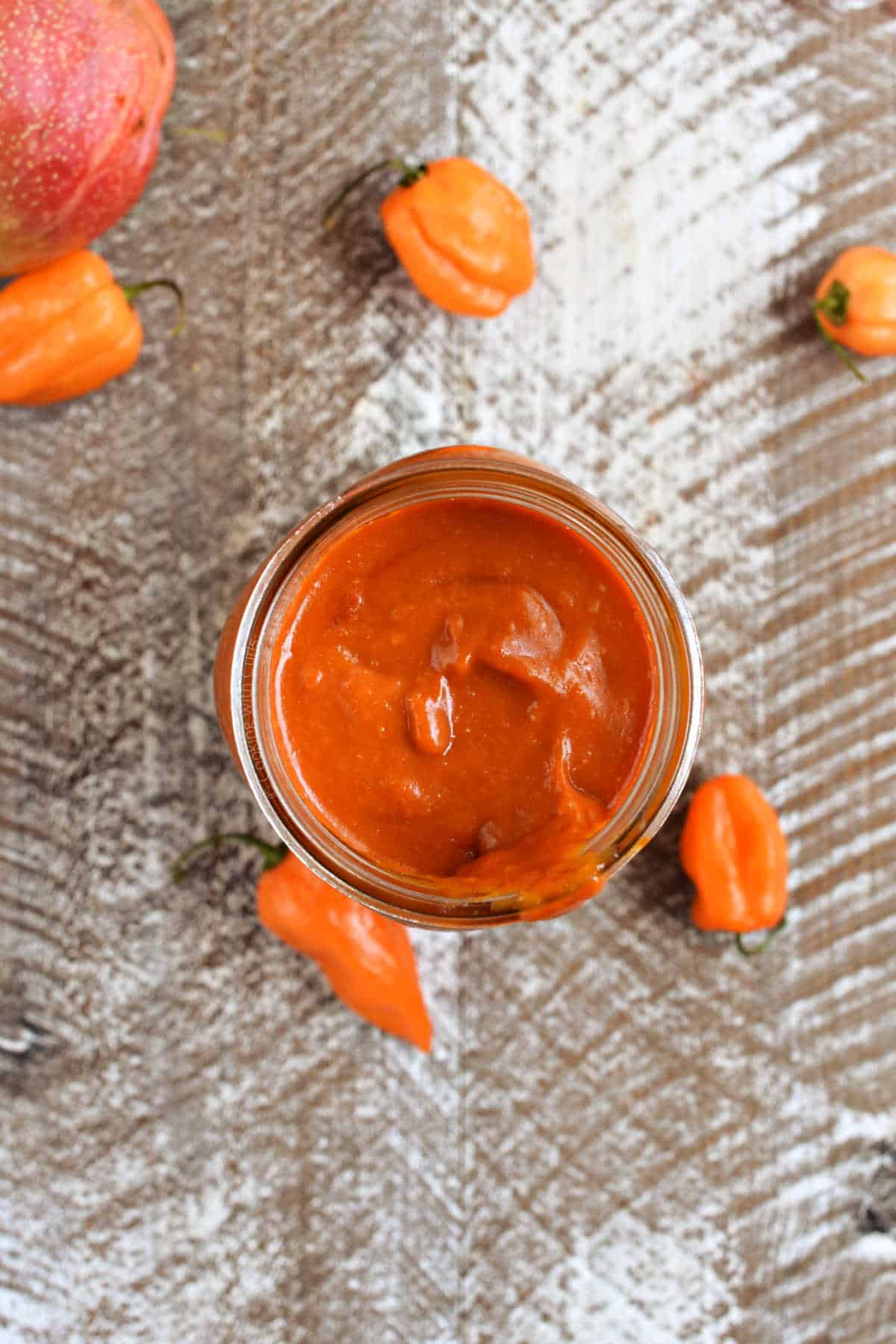 Reddish-orange bbq sauce in a mason jar with mango and peppers in the background.