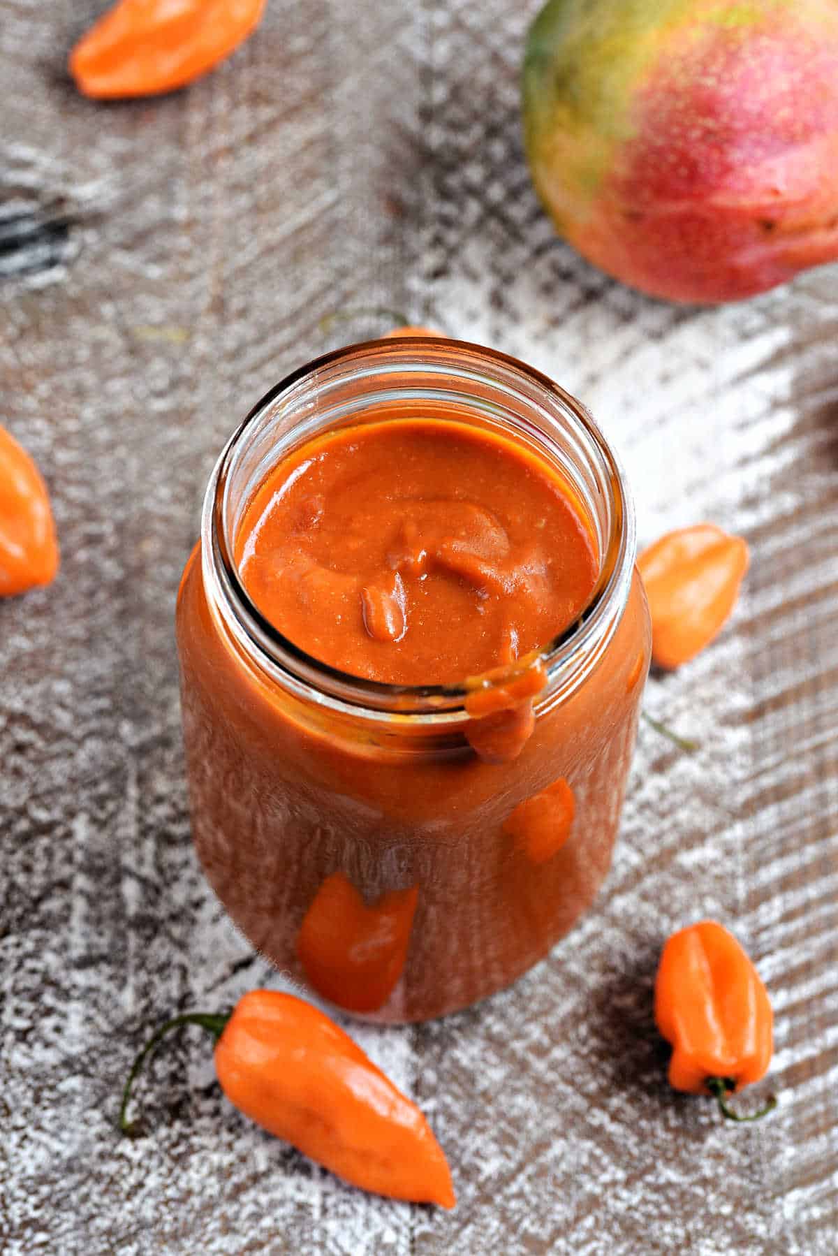 Deep reddish-orange barbecue sauce in a mason jar surrounded by habanero peppers.