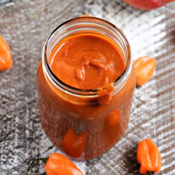 Deep reddish-orange barbecue sauce in a mason jar surrounded by habanero peppers.