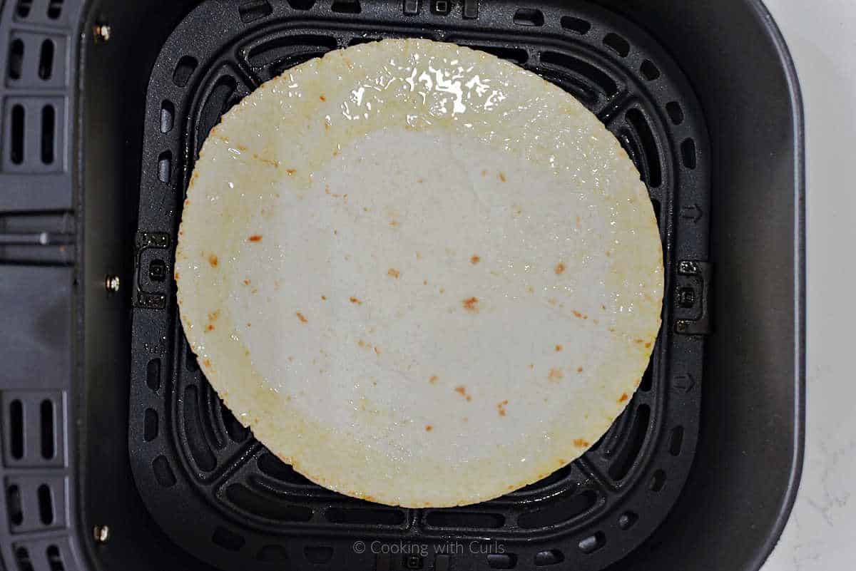 A flour tortilla with oil around the edges in an air fryer basket.