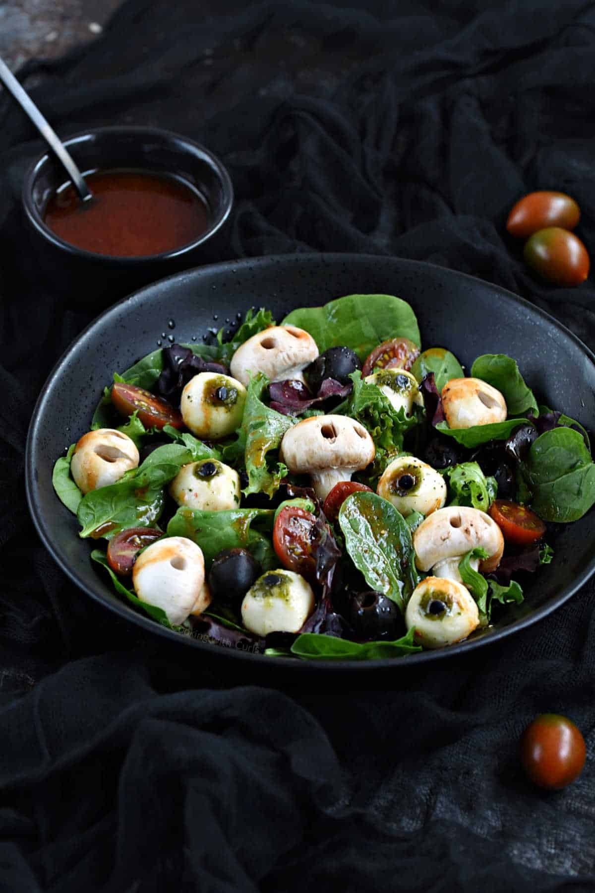 Mixed greens, mushroom skulls, mozzarella eyeballs, black olives, and blood red vinaigrette in a black bowl.