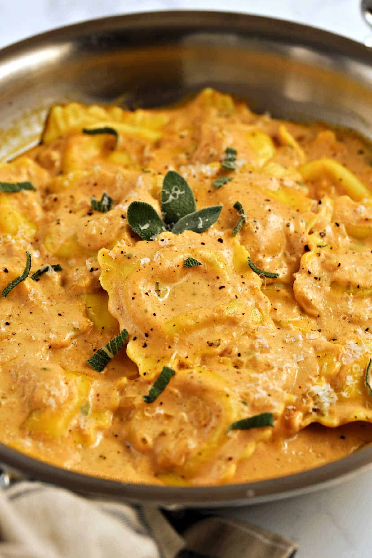 Ravioli covered in creamy pumpkin sauce, sprinkled with fresh sage leaves in a skillet.