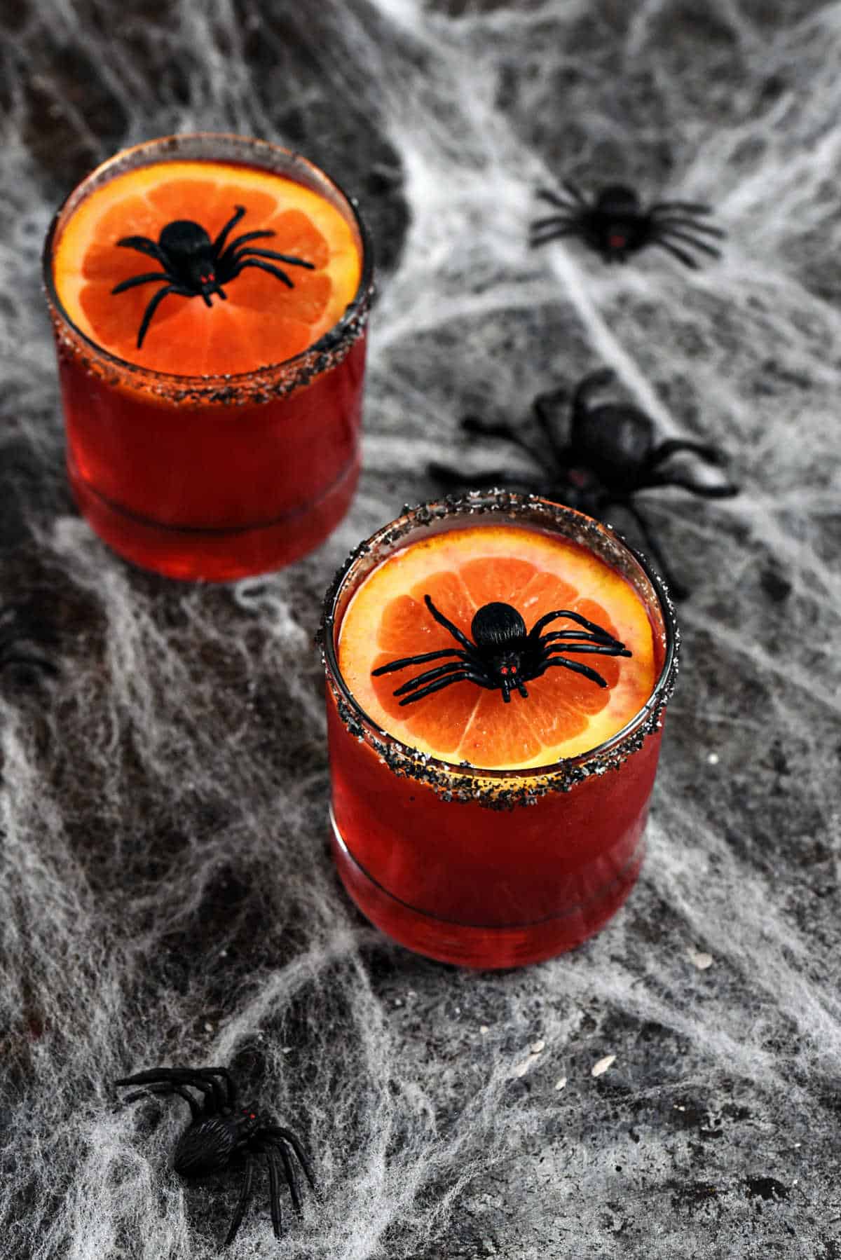 Red drink in short glasses topped with orange slice and plastic spider.